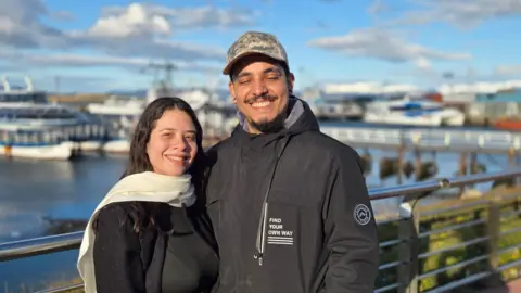 Matías Zibell / BBC News Mundo A couple in their twenties smile at the camera. Both are wearing black outdoor clothes. She has a white scarf. Behind them (blurred) is the harbour with a number of boats on the water. 