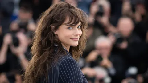 Getty Images Cara Delevingne wears a navy pinstripe jacket. She is looking over her shoulder and has brown wavy hair.