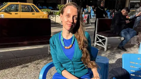 Getty Images Shelly Kittleson, who has long dark blonde hair and wears a blue dress with a turquiose cardigan, sits on a plastic chair in the sun in a Baghdad street 