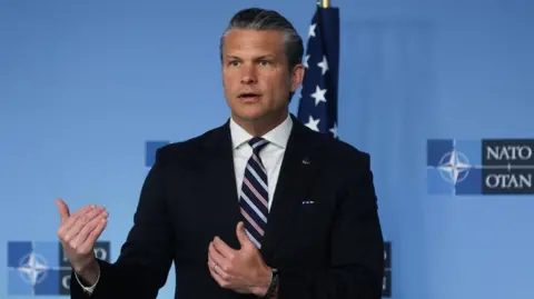 EPA Hegseth gestures with both of his hands as he stands in front of a Nato branded blue backdrop at a summit. He is wearing a dark blue suit, white shirt and white, blue and pink striped tie.