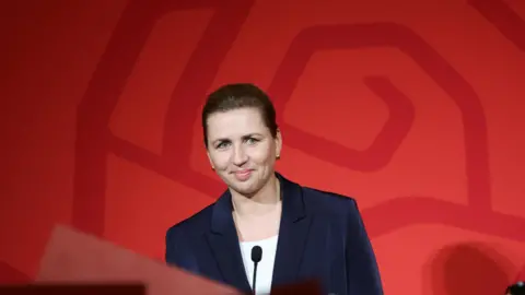 Reuters Mette Frederiksen, Denmark's Prime Minister, stands at a podium in front of a red background. She wears a blazer and is smiling slightly. 