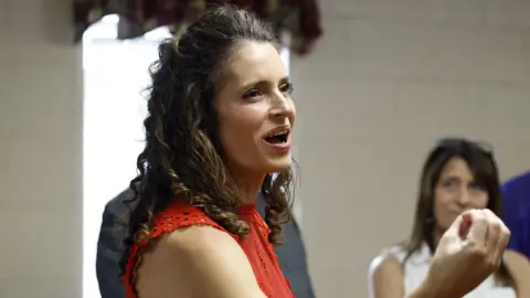 Tyler Kaufman/Getty Images Julia Letlow is seen talking to constituents which smiling and appearing to talk. She is turned to the side and is seen from the shoulder up. She is wearing a red top.