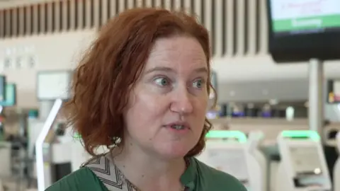 BBC Kate White, who has short red hair, speaks in a TV interview in front the check-in desks at Manchester Airport's departure lounge.
