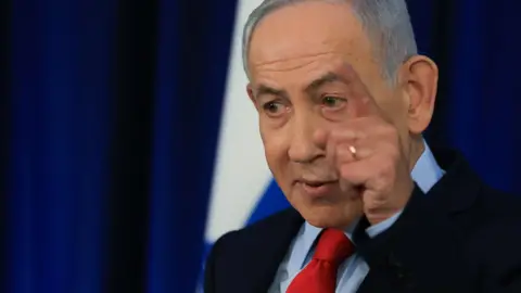 Reuters Benjamin Netanyahu stands speaking at a press conference. He wears a black suit jacket, blue shirt and red tie. One of his hands is raised, pointing his finger and he speaks. In the background, the Israeli flag is blurred.