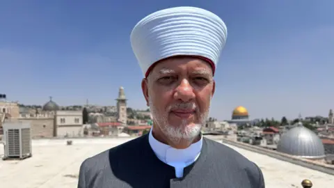 Maarten Lernout/BBC A bearded man in a grey tunic stands on a roof top in Jerusalem with the gold dome of the Al Aqsa mosque behind him 
