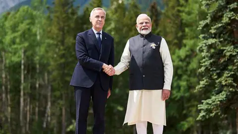 AFP via Getty Images Canadian Prime Minister Mark Carney shakes hands with Indian Prime Minister Narendra Modi. Both are facing forward, and are standing outside against a backdrop of trees. 