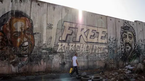 AFP File photo showing a man walking past portraits of Marwan Barghouti (L) and the late Yasser Arafat (R) on a section Israel's separation barrier, near the Qalandia checkpoint, in the occupied West Bank (22 September 2025)