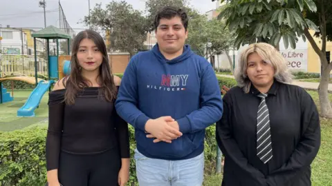 Consuelo dressed in black, next to Alvaro in a blue top and jeans and Cielo in a black top and tie. They are standing in front of an urban backdrop.