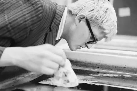 Getty Images David Hockney making a print in 1965. This is a black and white photo of the artist bending over his print and inspecting it closely.