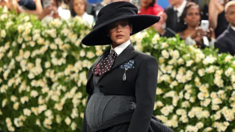 Getty Images Rihanna wears a black hat with a large brim and a stylised cropped suit jacket over a grey bodysuit emphasising a pregnancy bump 