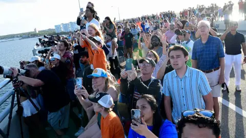 Gerardo Mora/Getty Images A group of people watches the launch, most holding phones or cameras up to the sky.
