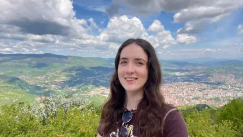 Em's Budget Travel Em, a young woman with dark hair. She is standing on a high road in front of a large green landscape with a town in the distance on the right.