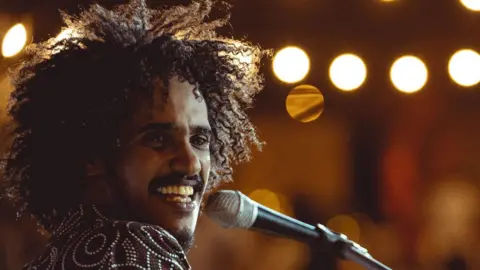 Diaa Talha A close-up of Ibrahem Mahmoud singing into a microphone. He has long curly hair and is smiling
