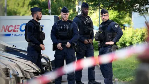 Getty Images Four uniformed French police stand in front of a row of cars, with a police tape in the foreground, near the scene of the shooting of three boys in the city of Nantes.