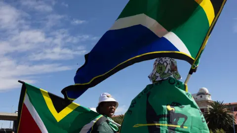 KIM LUDBROOK/EPA/Shutterstock Two people stand, waving South African flags, draped in other flags, and wearing hats, on a sunny day on the street.