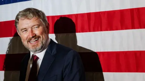 Getty Images Thomas Massie dressed in dark blue suit, red tie, with a grin on his face looking to the right. He's stood in front of a large USA flag