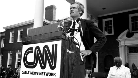 Getty Images Black & white photo of Ted Turner standing behind a lectern with a CNN logo at the launch event in Atlanta, Georgia, on 1 June 1980