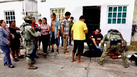 Darren Conway/BBC Julio César Vega and Héctor Torres tend to a man lying on the pavement while people look on. Two soldiers wearing uniform and helmets are also at the scene. 