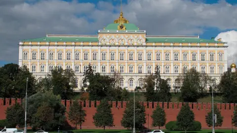 ullstein bild via Getty Images Picture of Kremlin Palace during the day. There is a blue sky and clouds. In front of the palace is a red brick wall and trees lined in front of it and behind it