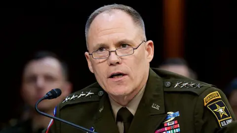 BBC LaNeve speaking into a small microphone that is on a stand in front of him. He wears small glasses and his military uniform which has several awards on it. The background is blurry but a man sits behind him