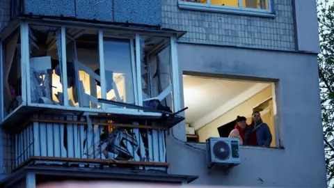 Reuters Two women look out of a window of an apartment block at the site of a Russian missile strike in Kyiv. Photo: 16 April 2026