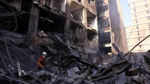 Reuters A rescue worker wearing a helmet and protective gear searches through rubble inside a heavily damaged multi-storey building with collapsed walls, twisted metal and debris.