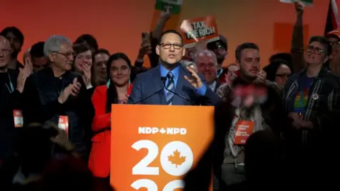 Reuters Avi Lewis speaking at a podium with crowd of supporters holding signs behind him