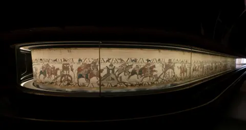 French Ministry of Culture The Bayeux tapestry presented in a curved display in a dark room.