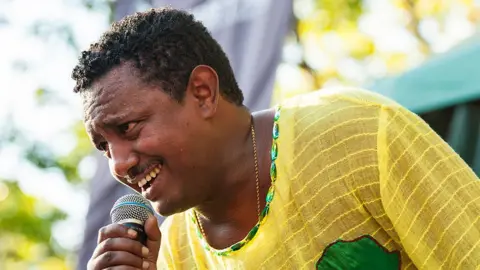 Getty Images A head and shoulders image of Teddy Afro who is leaning into a microphone in his right hand and is smiling. He is wearing a yellow T-shirt, and the top of the African continent in green can be seen sewed onto the shirt. 
