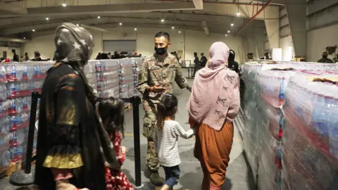 Getty Images A soldier guides Afghan Special Immigrants. Pictured here are some women and children seen from behind, into the dining facility, August 20, 2021 at Camp As Sayliyah, Qatar