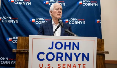 REUTERS/Joel Angel Juarez John Cornyn stands at a podium with red white and blue campaign signs. His expression is serious and he is looking into the distance.