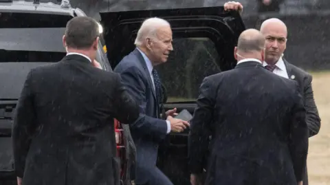 Getty Images Biden steps out of a black car in the rain surrounded by minders