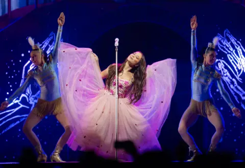 Getty Images Laufey lifts up the chiffon "wings" of her pink ballgown, while performing on stage at London's O2 Arena in March 2026