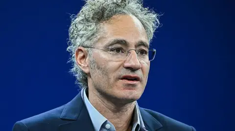 AFP via Getty Images Alex Karp, wearing a blue suit and shirt, speaks on a stage against a dark blue backdrop