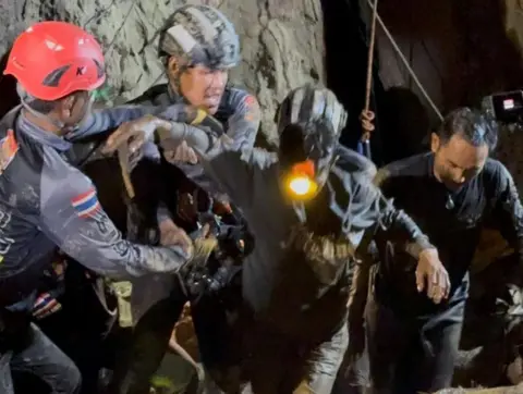 EPA Two rescuers in dark blue diving gear, with one wearing a black helmet and the other a red one, hold the right arm of the rescued man, who is dark muddy clothes and has a headlight on. His left arm is being held by another rescuer to his left as they all emerge from the cave in darkness