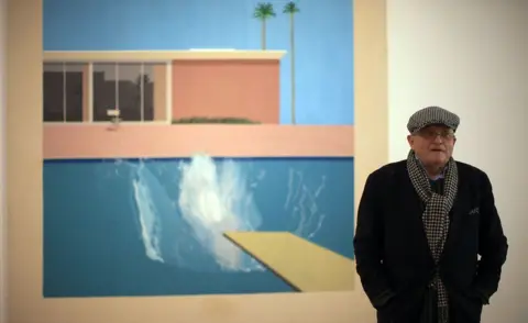 Getty Images David Hockney is shown in a gallery standing in front of his painting. The painting is of a big splash as someone has dived into a pool. Hockney is dressed in a cap and scarf even though it is indoors.