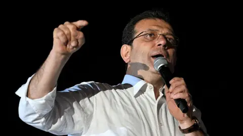 AFP via Getty Images Ekrem Imamoglu gives a speech following municipal elections in Istanbul in 2024. He is holding a microphone in one hand while his other is pointed towards his supporters. He has glasses on and is wearing a white shirt.