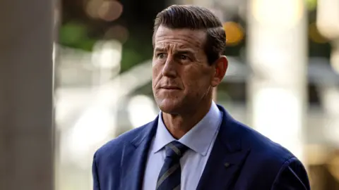 Sam Mooy/The Sydney Morning Herald via Getty Images A front-on shot of Roberts-Smith looking pensively away from camera while wearing a suit and tie with strong background bokeh, as he walks along a street while arriving at the Federal Court in Sydney in May 2025.