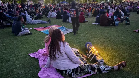 A group of people, some wearing pink, sit on the grass and light candle lights during the vigil for the little girl's death.