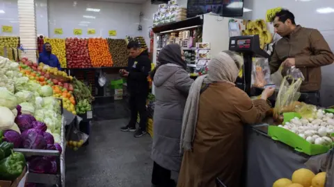 EPA Scene inside a fruit and vegetable store, where two women are buying an item from a man at the till and two other men can be seen in the background.