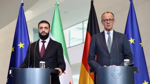 Reuters German Chancellor Friedrich Merz and Syrian President Ahmed al-Sharaa wearing suits and standing at podiums, in front of flags, hold a press conference at the Chancellery in Berlin, Germany