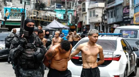 ANTONIO LACERDA/EPA/Shutterstock Rio de Janeiro Police officers transfer a group of people detained during an operation in Rio de Janeiro, Brazil, 28 October 2025. The officer is holding a weapon and is wearing a balaclava. The suspects are stripped down to their waist. They both have tattoos. 