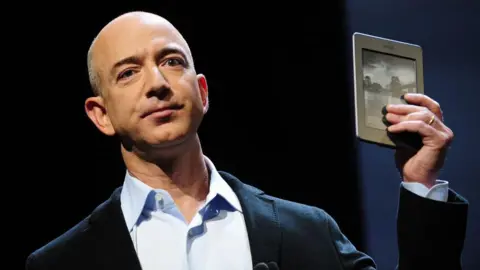 AFP via Getty Images Amazon founder Jeff Bezos, in dark blue blazer with light blue shirt, posing on-stage in 2011 with a Kindle Touch