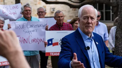 Getty Images Senator John Cornyn, a Republican from Texas, speaks to members of the media after casting his ballot at a polling location inside the Circle C Community Center on the first day of early voting in the Texas runoff election in Austin, Texas. Several people stand behind him carrying posters supporting his run.