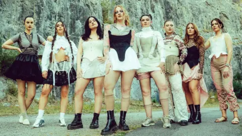 Claire Loughran Eight women stand in a line outdoors in front of a rocky cliff, wearing coordinated, fashion-forward cream and black outfits with boots and layered textures, posing on a roadside against a natural stone backdrop.
