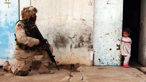 Stefan Zalkin/EPA-EFE A US soldier guards a street during a patrol, he kneels on the concrete floor with his gun pointing down. He wears a camoflauge uniform. A young girl stands around the corner of a building looking at him.