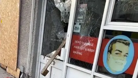 Steve Aiken A window of a barber shop is smashed. Some boarding has been put up next to the window. Wood is hanging out of the door. 