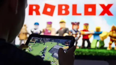 AFP via Getty Images A young person plays Roblox in front of a larger screen showing the Roblox logo.