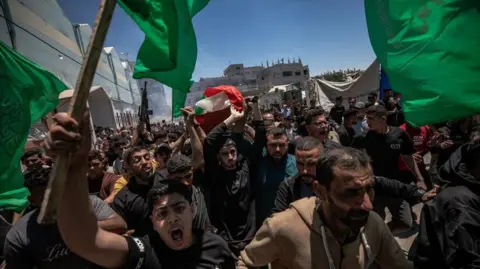 EPA/Shutterstock Dozens of people taking part in a funeral procession through Gaza City, some waving green flags and one holding a gun aloft 
