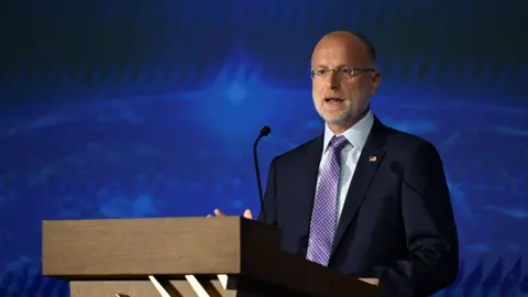 Reuters FCC chairman Brendan Carr speaking at a wooden podium wearing a navy suit, white shirt and red and blue checked tie.